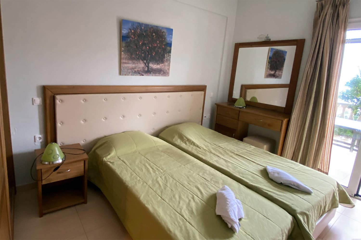 ein Schlafzimmer mit einem Doppelbett und hellgrünen Tagesdecken. An der Wand hängt ein Spiegel und ein Bild mit einem Olivenbaum