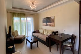 Olive Tree Wohn- & Essbereich ein Sideboard auf dem ein Flachbild-Smart-TV steht. Daneben ein Sessel und in der Mitte ein kleiner Wohnzimmertisch, dahinter ein Sofa. An der Wand hängt ein Bild von einem riesigen Olivenbaum