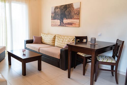 ein 2er Sofa und ein Esstisch an dem zwei Stühle stehen. An der Wand hängt ein großes Bild mit einem riesigen Olivenbaum